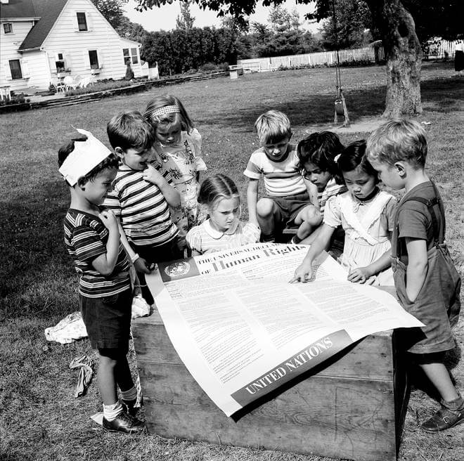 Photo en noir et blanc de 1950. Des enfants des membres du personnel des Nations unies regardent attentivement une grande affiche de la Déclaration universelle des droits de l'Homme.