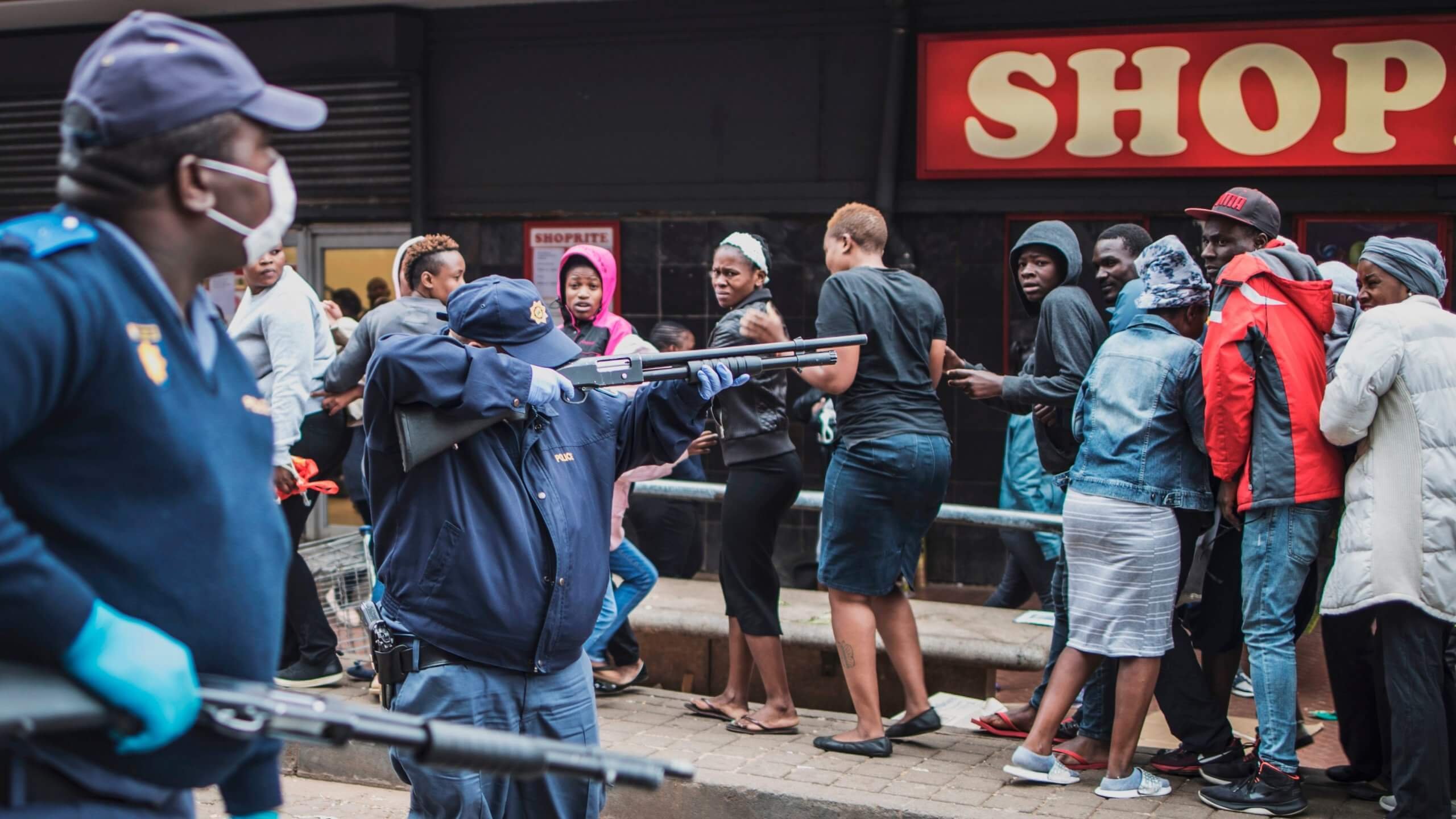 TOPSHOT - Un policier sud-africain pointe son fusil à pompe pour disperser une foule d'acheteurs à Yeoville, Johannesburg, le 28 mars 2020, alors qu'il tente de faire respecter une distance de sécurité à l'extérieur d'un supermarché dans l'inquiétude de la propagation du coronavirus COVID-19. - Le 27 mars 2020, l'Afrique du Sud s'est retrouvée sous le coup d'un embargo national, rejoignant ainsi d'autres pays africains qui ont imposé des couvre-feux et des fermetures strictes pour tenter de stopper la propagation du coronavirus COVID-19 sur le continent.
